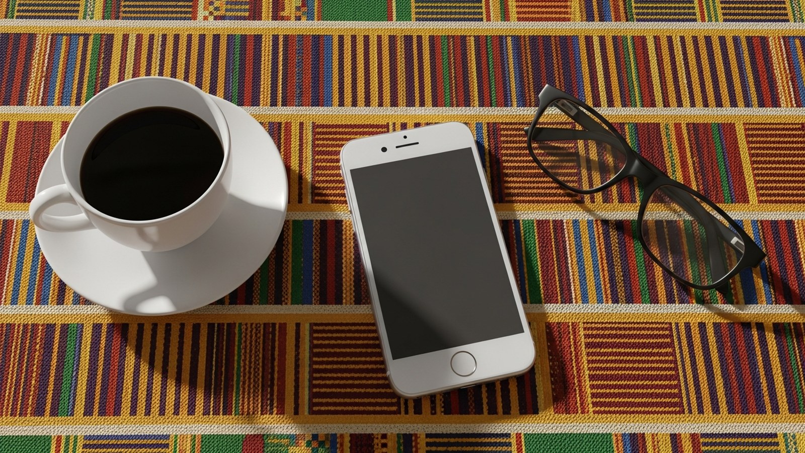 Overhead view of black coffee, a face-down phone and reading glasses on a kente-pattern cloth in morning light